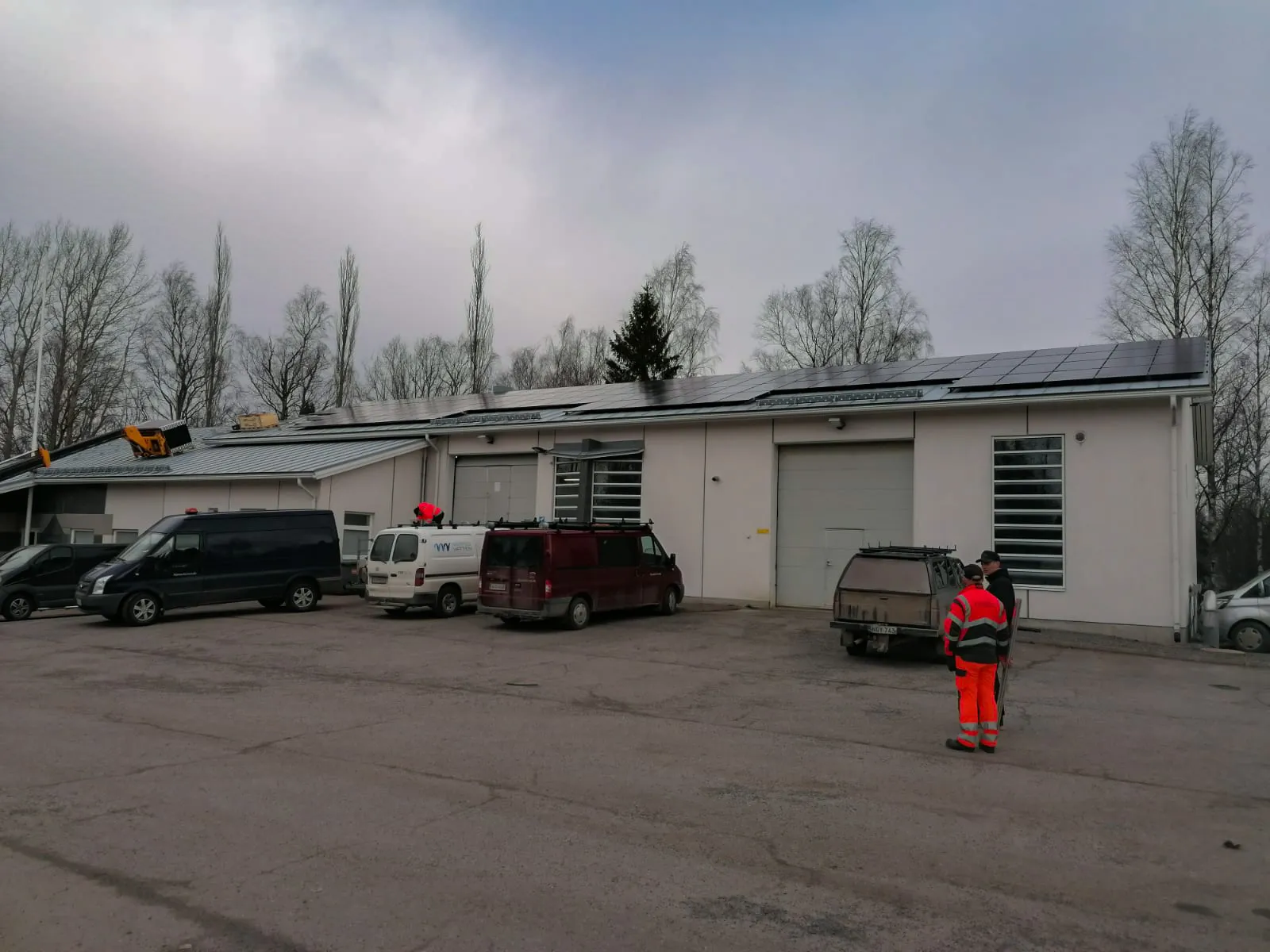 Rakennuksen katolla on aurinkopaneeleita ja pihalla useita pakettiautoja sekä työntekijöitä.